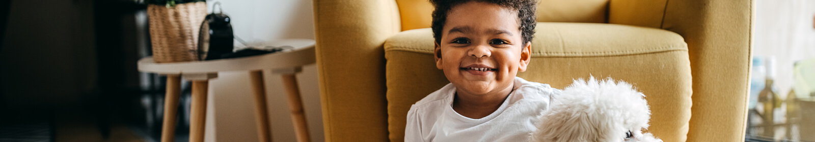 A toddler smiling in family living room while holding their dog.