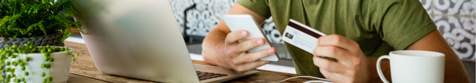 Person holding debit card and smart phone while sitting at home desk in front of laptop.